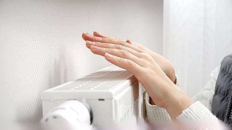 Woman in sweater and scarf warming her hands at heating radiator on the cold day. Concept of energy crisis, high bills, broken heating system, economy and saving money on monthly utility payments.