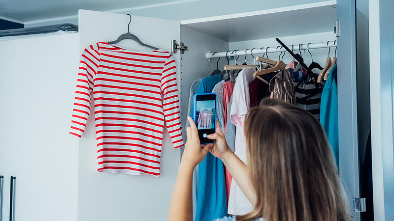 Woman taking photo of the shirt using her smartphone for selling or donating her clothes. Decluttering , Sorting Clothes, And Cleaning Up wardrobe. Reuse, second-hand concept. Conscious consumer