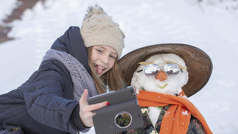 Adorable young girl is taking pictures of selfie with a snowman in beautiful winter park. Winter activities for children.