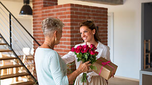 Happy adult daughter bringing gift and bouquet to senior mother indoors at home.
