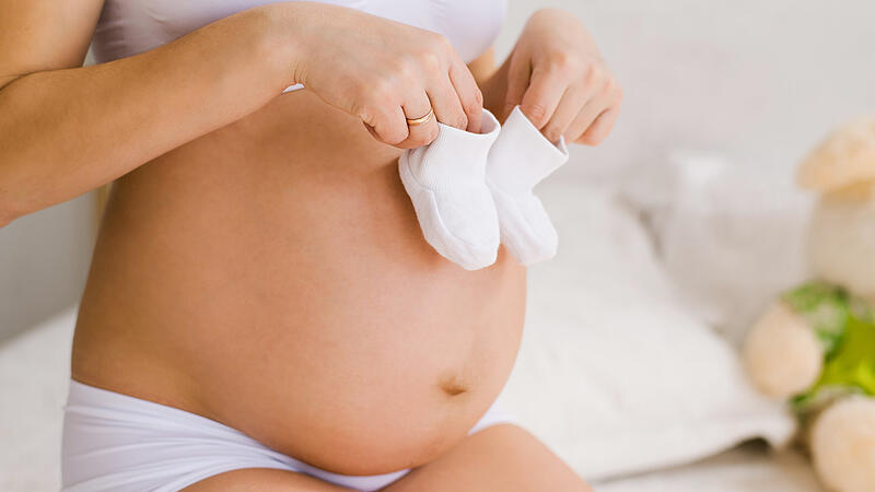 A woman with a big bare tummy is sitting on the bed holding baby booties in her hands. Pregnant lady strokes her belly. Tiny socks on a big belly