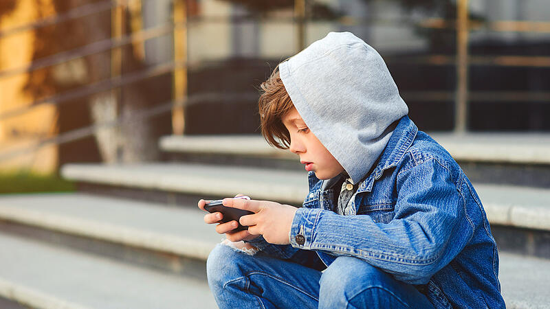 Boy use phone and plays games. Kid in the hood sitting on the stairs. Kids addicted online games and cartoons. Schoolboy plays on smartphone after school.