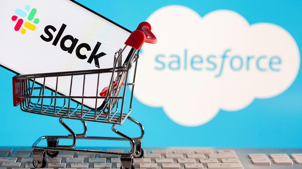 Smartphone with Slack logo stands in a shopping cart on a keyboard in front of displayed Salesforce logo in this picture illustration