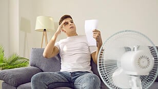Sweaty man feels bad because of terrible heat sitting at home in front of fan during hot summer days